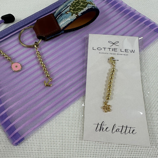 Gold beaded charm on a card with 'Lottie Lew' branding, next to a keychain and fabric strip.